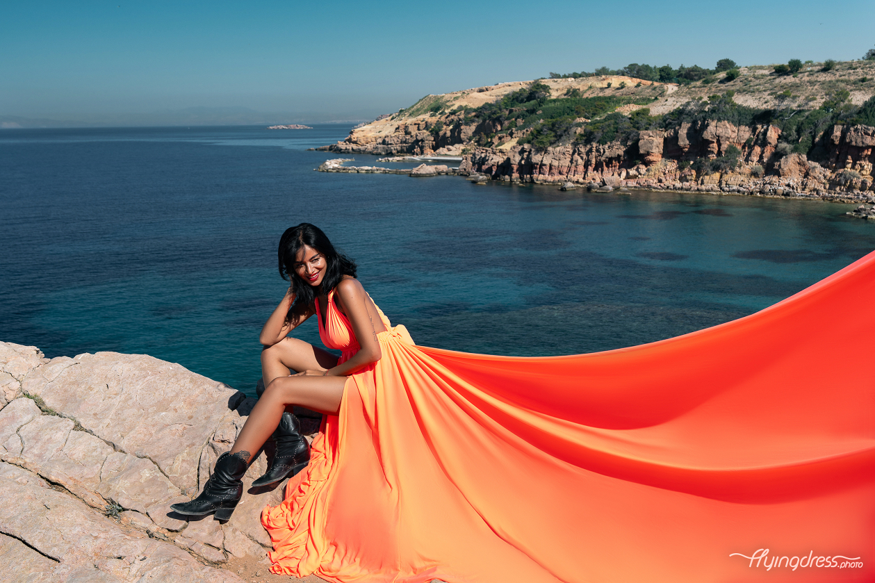 A woman in a vibrant orange flowing dress and black boots sits gracefully on rocky terrain with the clear blue waters and scenic coastline of Kavouri, Athens in the background.